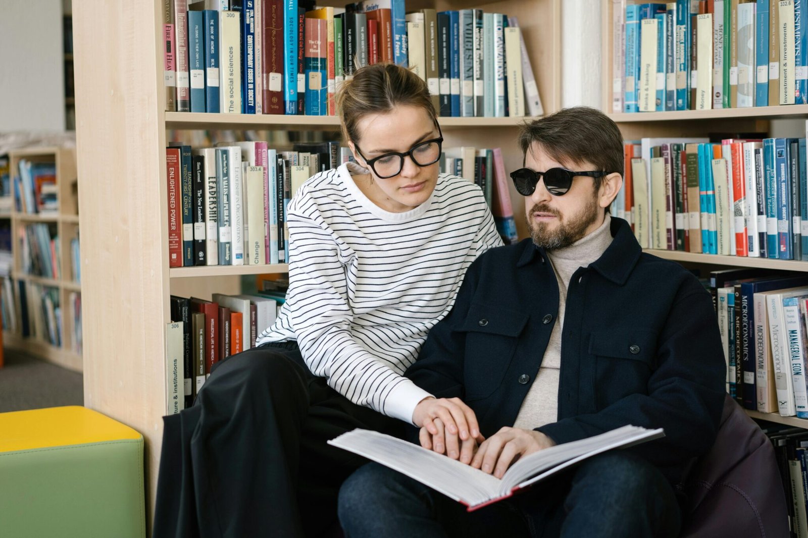 A woman helps a visually impaired man read a braille book in the library.